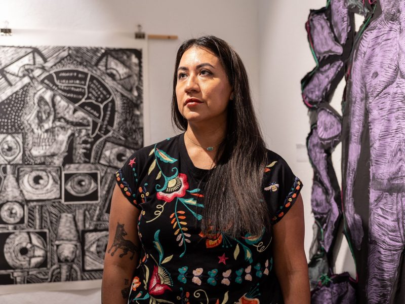 Artist Celeste de Luna poses in front of two of her artworks on display inside of Guadalupe Bookstore as part of the Life and Death on the Border 1910-1920 traveling exhibit created by Refusing to Forget.