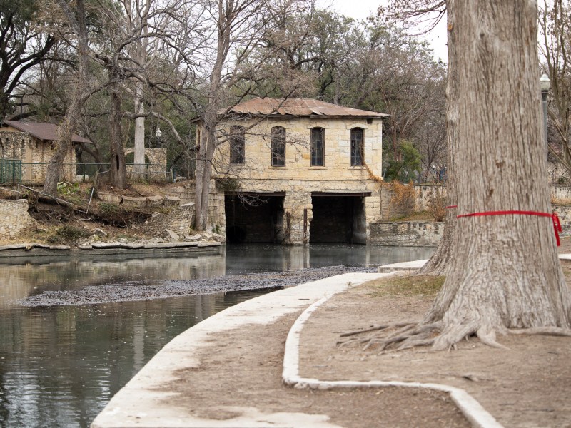 The historic pump house and Lambert Beach at Brackenridge Park are flanked by retaining walls that are crumbling due to encroaching tree roots and other factors. Some trees mislabeled by activists to be removed sit across the river.